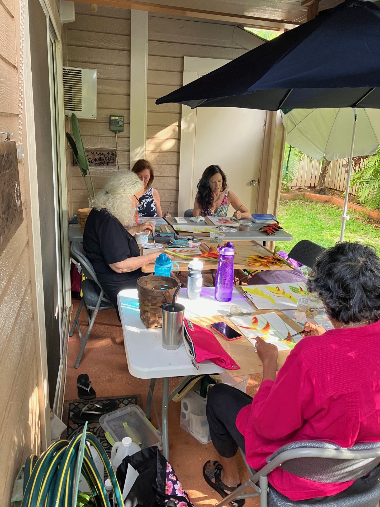 Group of adult students painting tropical flowers outdoors under a patio umbrella at Steinberg Art School